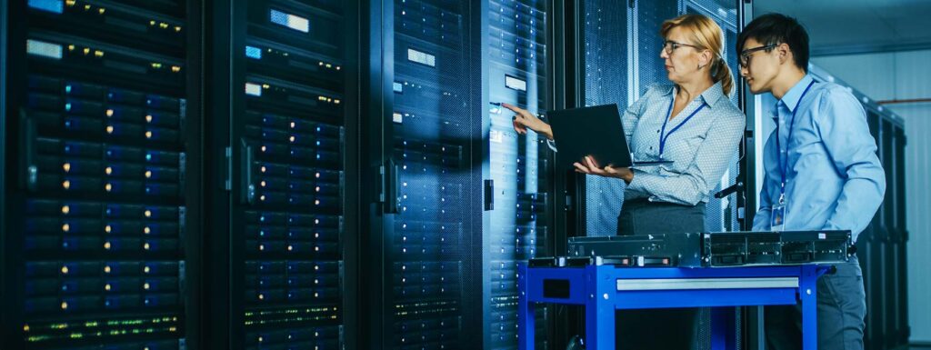 Two IT professionals reviewing data on a laptop inside a data center, supporting infrastructure management and technology operations
